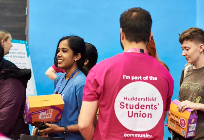 Person in a pink "Huddersfield Students' Union" shirt handing out boxes to other individuals at an indoor event.