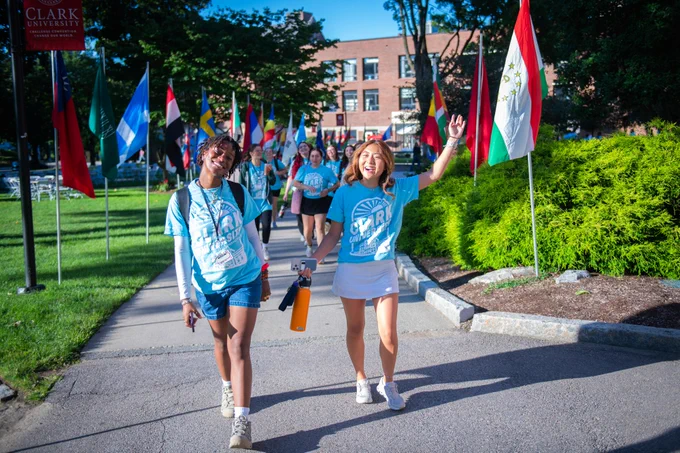 Two individuals in "Clark University Orientation" T-shirts joyfully walking on a path lined with international flags, with campus buildings in the background.
