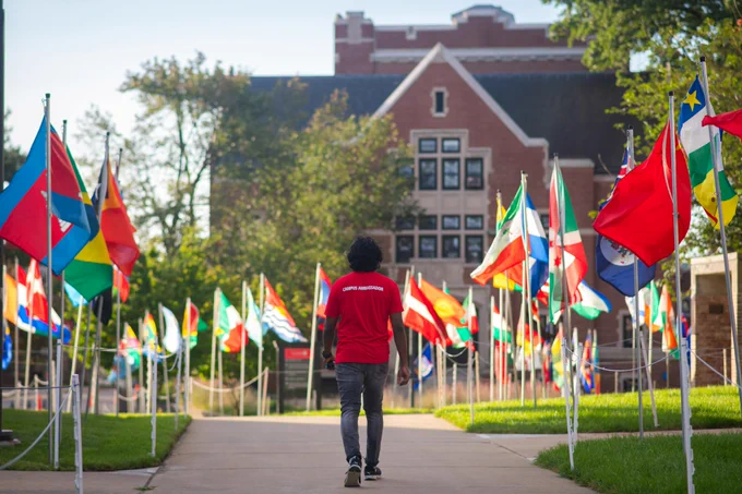 Person in a red shirt walking along a pathway lined with various international flags, with a campus building in the background.
