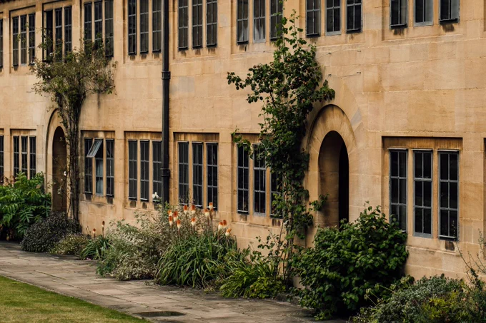 The staircases of Lower Quad at Nuffield College. Many multi-paned windows over two floors in a Cotswold stone building, intersperse with curved doorway arched. In front of the windows are mature plants, a paved walkway and a hint of lawn. Photo by Tom Weller.