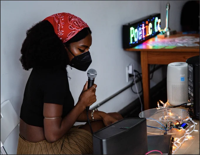 Photo of a block party assistant, holding a microphone. She has black skin, a red bandana on her hair and is masked.