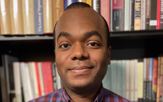 A Black man stands in front of a bookcase.