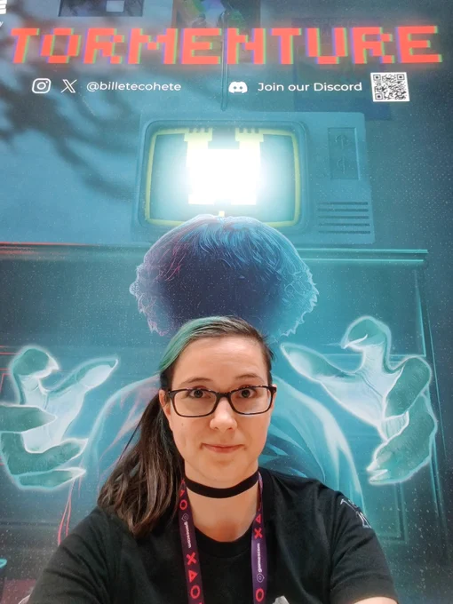 Key art of the upcoming game Tormenture showing a box sitting in front of a TV with ghost hands attempting to grab him. 2DKiri is sitting in front of the poster with wide eyes.