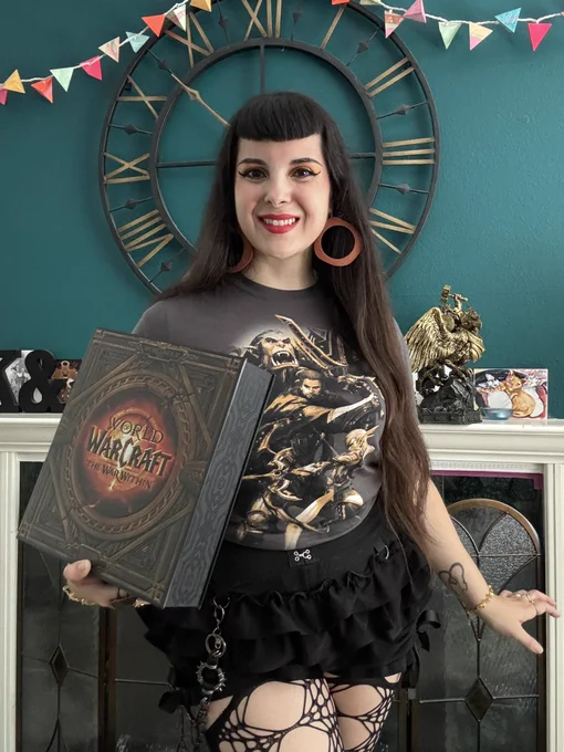 Kristy stands in front of her fireplace set into a turquoise wall with a metal clock framing her head. She has long dark hair and is wearing a gray and gold tee shirt featuring the WoW characters Thrall, Anduin, and Alleria. She is smiling and holding a WoW War Within collector’s edition box.