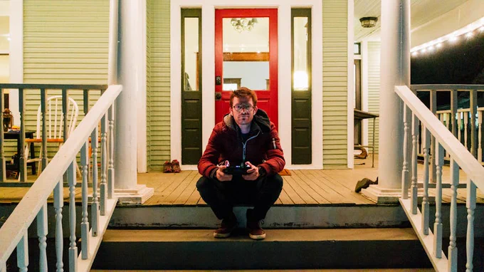 Full body male in warm clothes and round eyeglasses browsing internet on tablet and looking at camera while sitting house porch and chilling