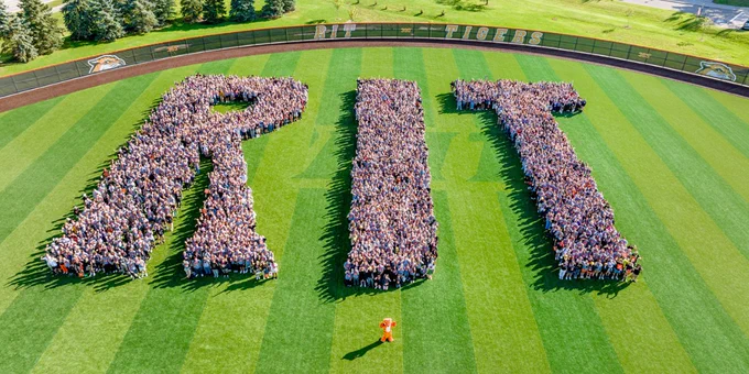 Students stand in groups forming the letters R, I, and T on a baseball field with the tiger mascot Ritchie.