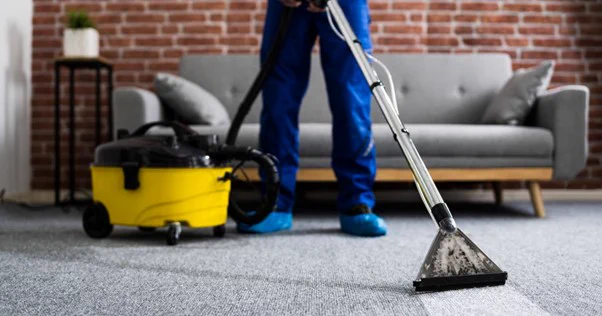 Image of a person wearing blue overalls and shoes, and hoovering a grey carpet. There is a grey sofa, black side table, and brick wall in the background.