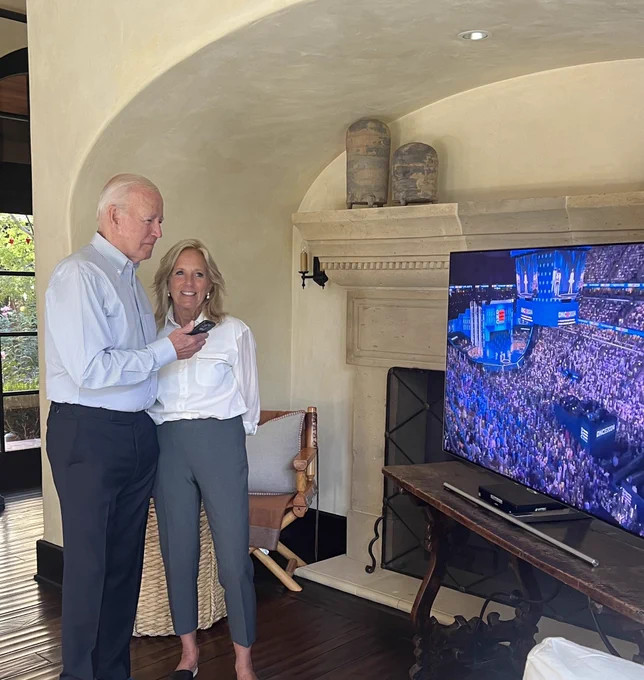 President Joe Biden and First Lady Jill Biden speak to Vice President Kamala Harris by phone while standing to the left of a TV playing a stream of the Democratic National Convention.