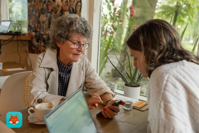 "Twee vrouwen zitten aan een tafel in een café. Een oudere vrouw met grijs haar en een bril toont iets op haar smartphone aan een jongere vrouw. Op de tafel staat een kopje koffie en een open laptop. Op de achtergrond zijn planten en een raam met uitzicht op groen te zien."