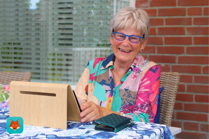 "Een oudere vrouw met kort, lichtgrijs haar en gekleed in kleurrijke kleding zit lachend aan een tafel. Ze houdt een tablet vast die in een houten standaard staat, met een smartphone en een portemonnee die op de tafel liggen. Op de achtergrond zijn een bakstenen muur en een raam met jaloezieën te zien."