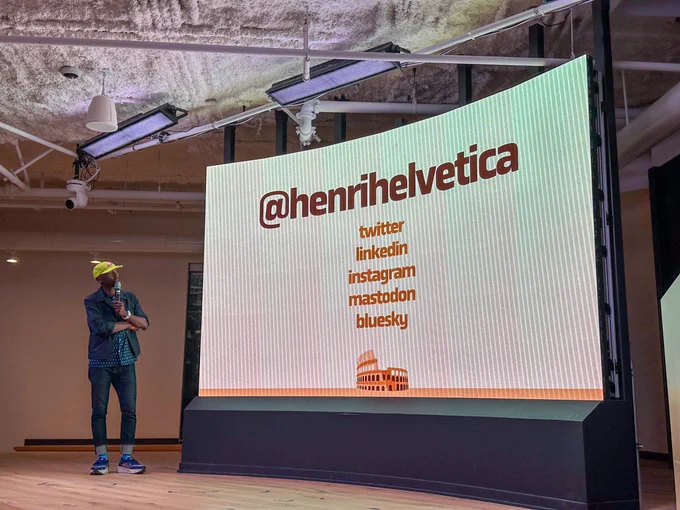 a person in a yellow cap, jeans and a jacket holding a mic on a stage, stand next to a large screen that reads "@henrihelvetica. Twitter, LinkedIn, Instagram, Mastodon, Bluesky"