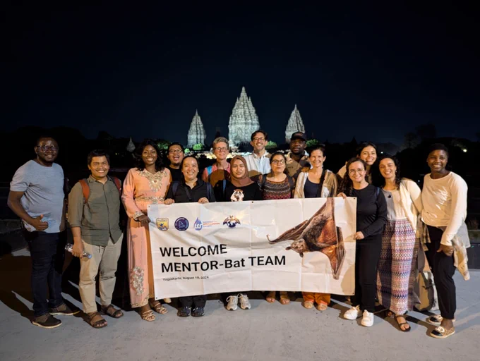 A group of about 20 people standing in front of a lit up building at nighttime, holding a banner that reads "Welcome MENTOR-Bat Team." 
