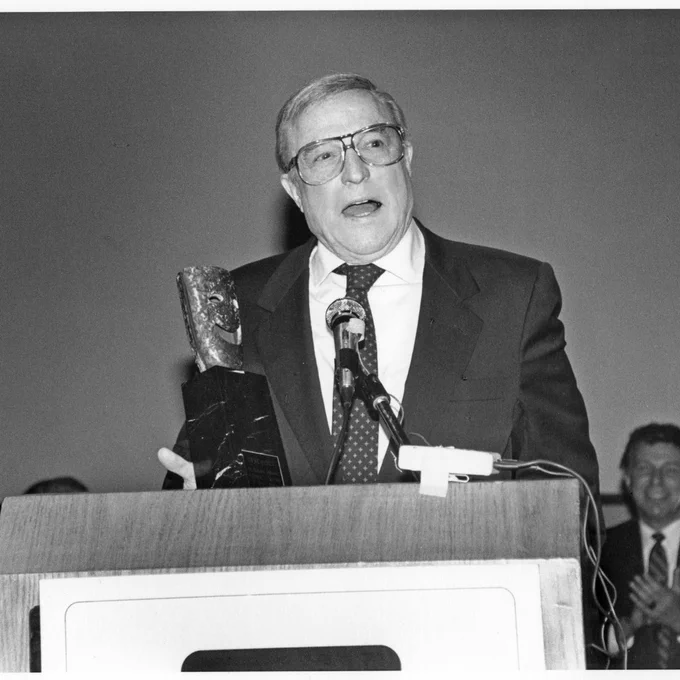 Gene Kelly standing at a podium with a microphone, his head tilted slightly to the right as he talks, looking off into the distance. His SAG Life Achievement Award statuette sits on the podium in front of him.