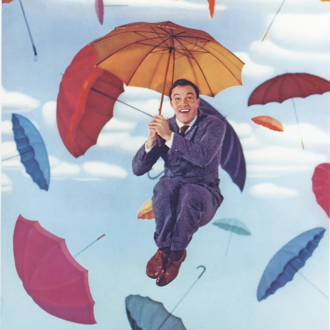 Gene Kelly, on a background of a blue sky with clouds and various colored umbrellas falling, holds an orange umbrella with both hands, his knees tucked to his stomach in a jumping motion.