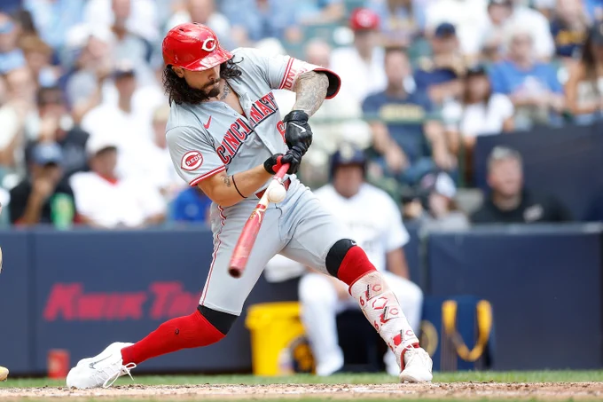 Pictured: Jonathan India mid swing, making contact with a baseball. He wears a road gray Reds uniform with a red batting helmet.