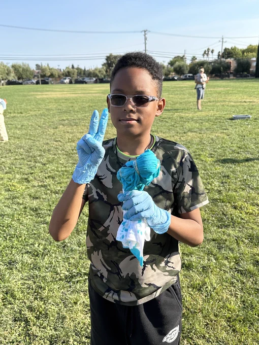 A student holds up a blue and white tie-dye shirt; he is wearing shades, a camo shirt and blue gloves with black shorts while standing on the grass at John McCandless