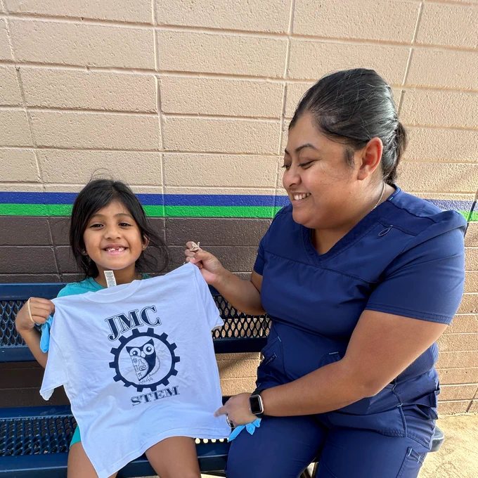 A mother and student sit on a blue bench as they hold up a white t-shirt with a John McCandless Owl logo at the words "JMC STEM"