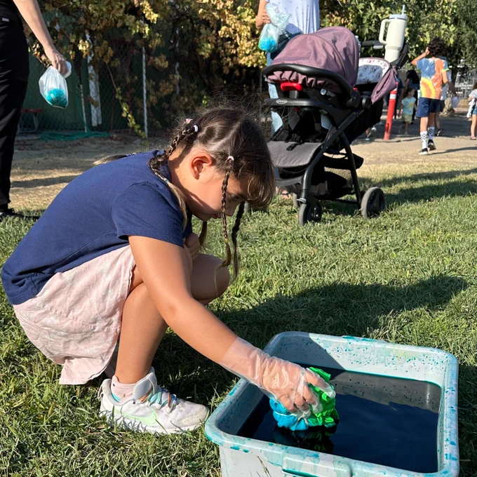 A girl kneels on the grass to soak a t-shirt in blue and green water coloring
