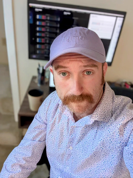 Selfie of Joe sitting in front of his home office desk wearing a light purple baseball cap embroidered with the Figma wordmark