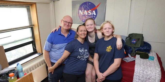 Four individuals smiling in an RPI dorm room with a NASA poster on the wall behind them. They are gathered closely together in front of a bed.
