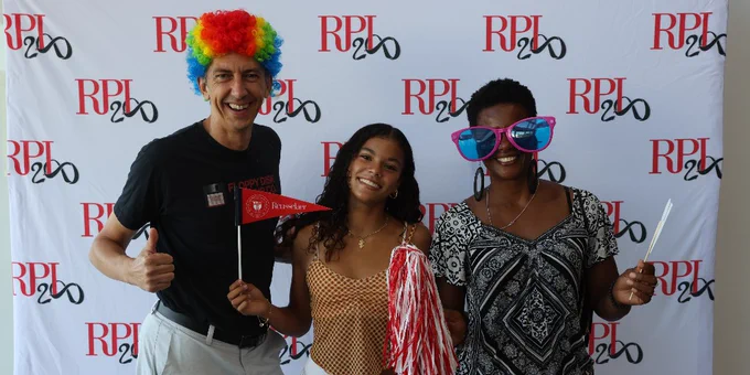 Three people posing with props in front of a backdrop with the RPI logo repeated.