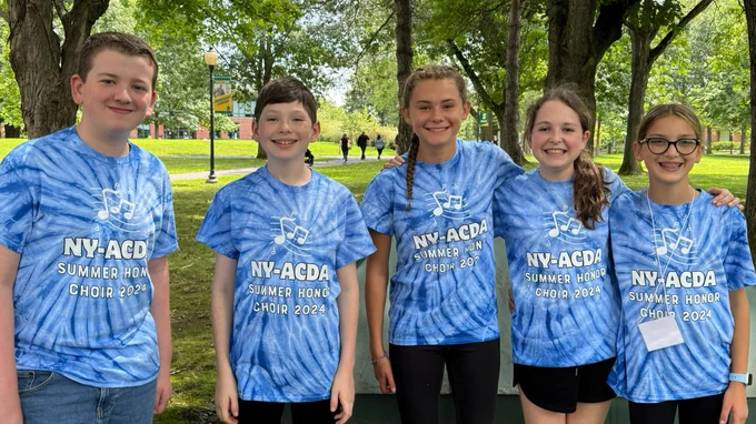 Five smiling middle school kids in blue shirts standing in front of trees.