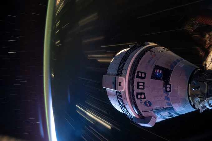 Boeing's Starliner spacecraft, docked to the International Space Station, zooms in orbit above the Earth. Starliner, a white cone with a cylindrical base, is attached to hardware just barely visible on the right side of the image. The Earth at night, with cities and other light sources as long streaks, takes up most of the background. Credit: NASA/Matthew Dominick