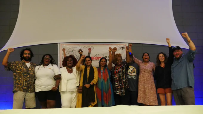 A group of brown, Black, and white people standing in front of a poster with fists raised, mouths open. 