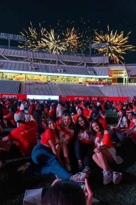 Group of people wearing University of Houston shirts sitting on the TDECU Stadium field, smiling for a photo, with fireworks visible in the sky above the stadium.