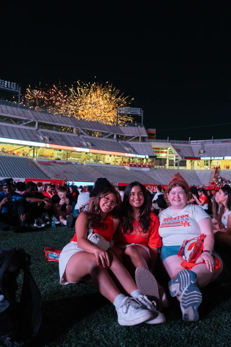 Three students seated on the field at TDECU Stadium, smiling, with fireworks bursting in the night sky above them. They are dressed in casual attire, prominently featuring red and white colors.
