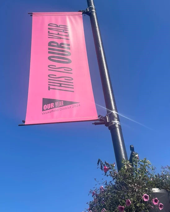 A bright pink banner with black text reading 'THIS IS OUR YEAR' is hanging from a pole against a clear blue sky. The banner is promoting 'OUR YEAR Wakefield District 2024'. 