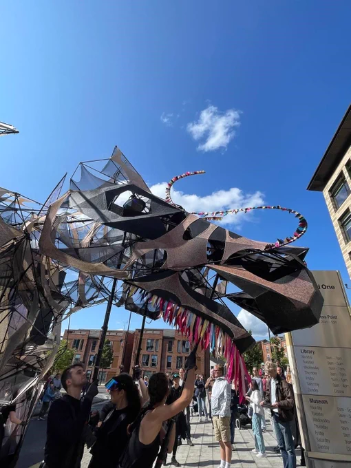 A group of people are interacting with The Hatchling dragon puppet. The structure is made of mesh-like material with colourful ribbons and geometric shapes, and the sky is clear and blue.