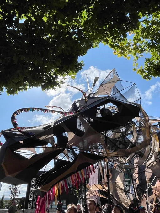 The Hatchling dragon large-scale puppet outdoors under trees, with people below looking up at it. The sky is bright and blue with some clouds.