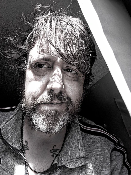 photo of odspoor/mitch in black and white, peering out his window with shaggy hair, big eyes, and a habs sweater unzipped to reveal two udjat tattoos on his chest from his wilder days lol. 