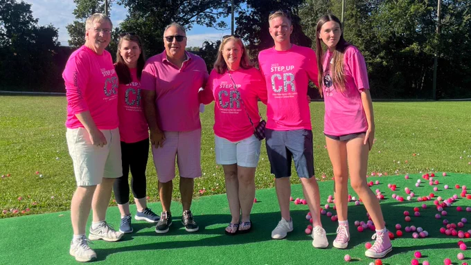 Six people in pink shirts standing on a driving range with pink golf balls on the ground.