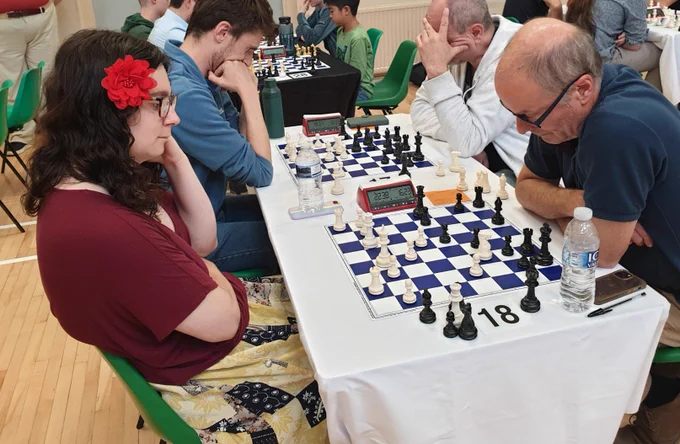 A picture of two chess players (me on the left), with the camera angled from the side of the chess board so both players are in profile. There are more chess sets and players in the background.