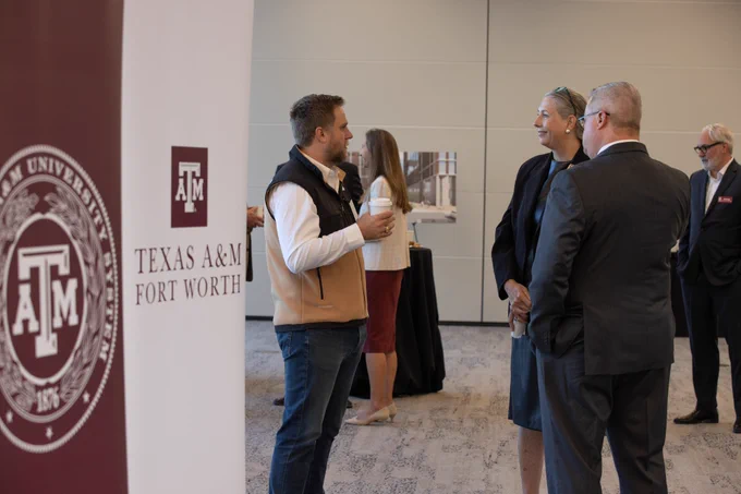 Individuals talk outside of an event space beside a Texas A&M University System banner
