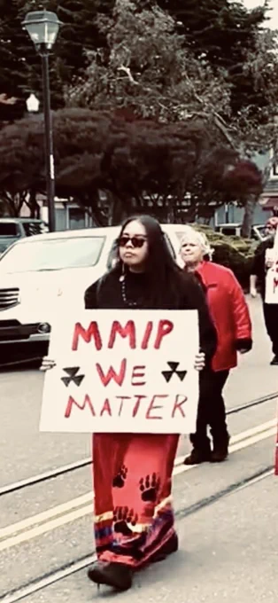 FBQ holding a Missing and Murdered Indigenous Peoples “we matter” sign during a MMIP awareness walk. FBQ is wearing a red ribbon skirt with bear paws and a black sweat shirt and sunglasses.