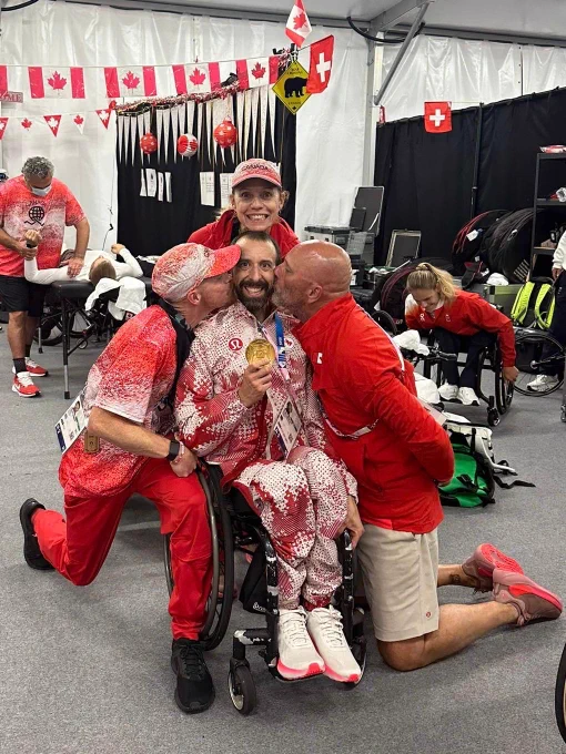 A man in a wheelchair smiling with a gold medal from the Paralympics Games. Two other men are kissing his cheeks and a woman is resting her head in his