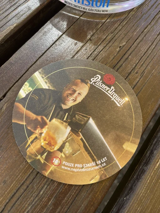 A drink coaster on a wood table. The picture on the coaster is off a guy that looks like a Czech Innkeeper Hatch (according to fellow fans who see it). This person is holding a mug of beer and smiling, and appears to be standing at a bar.