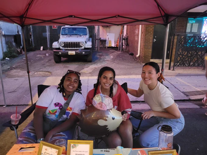 Photo of RA Abria, RA Chloe, and Lab Manager Alicia posing at our recruitment table at Friday Night Lights. Chloe is also holding a hedgehog balloon who is wearing a brain hat! 
