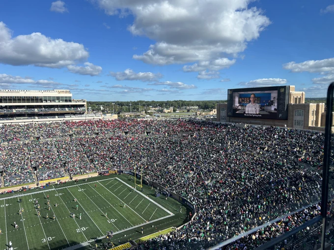 Aerial view of a crowded Notre Dame Stadium during a daytime football game, with clear skies and a large video scoreboard displaying an image of economist Patrick Turner.