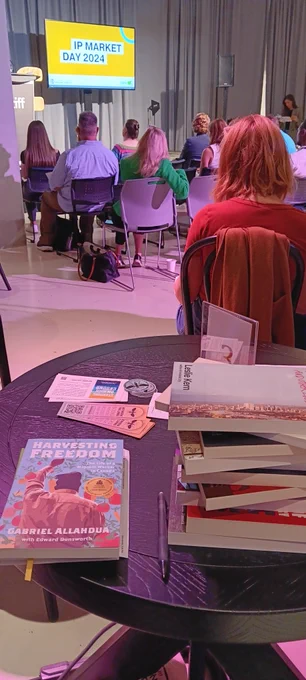 Photo of people at IP Market Day. In background: a yellow slide with the words "IP Market Day 2024" on it. People are sitting facing the screen. In the foreground is a round wooden table with a sampling of BTL books on it.