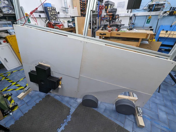 The assembly takes shape.  This image is in the hackspace workshop area. There are lot of benches and tools in the background. In the foreground is the Bosch strut aluminium spar frame. The bottom hafl, up to where the screen will be installed is cald in white panels. There is a cutout for the joypad and the large black joupad is patrialliy installed. on the blue tiled floor, to the bottom right of the fram and panels the two round buttons sit on the floor ready to be installed.