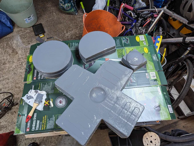 The joypad button, which is about 500mm square, and the two round buttons sit on an old cardboard box in a garage and are painted in grey primer. A paintbrush loaded with grey paint sits to one side. In the background is an assortment of bicycles, trugs and other domestic garage contents.
