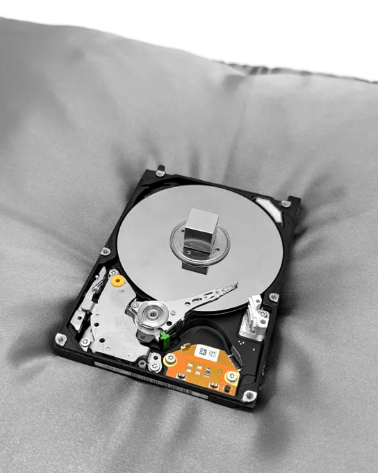 An exposed computer hard drive (HDD Toshiba MK2109MAT 2GB 5.4K IDE 2.5") rests on a silver satin royal pillow. The pillow has four tassels on the corners made from computer cables and connectors.