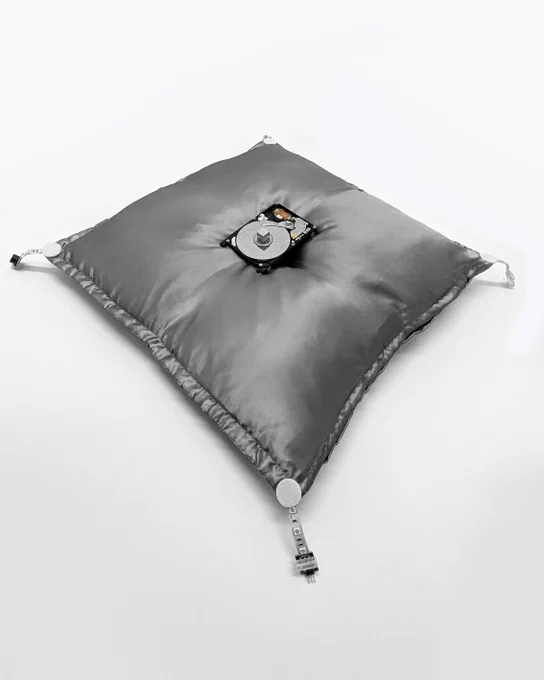 An exposed computer hard drive (HDD Toshiba MK2109MAT 2GB 5.4K IDE 2.5") rests on a silver satin royal pillow. The pillow has four tassels on the corners made from computer cables and connectors.