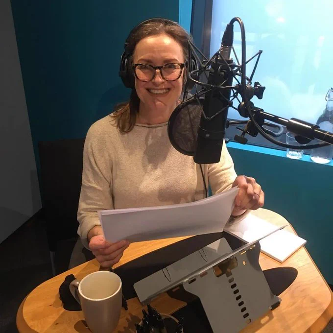 Heather wears a beige top and tortoiseshell glasses and sits in front of a recording microphone wearing headphones and holding a script