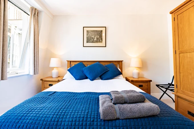 Double bed with white linen and navy cushions and thow. Wooden wardobe to the left hand side anf grey towels stacked on the bed