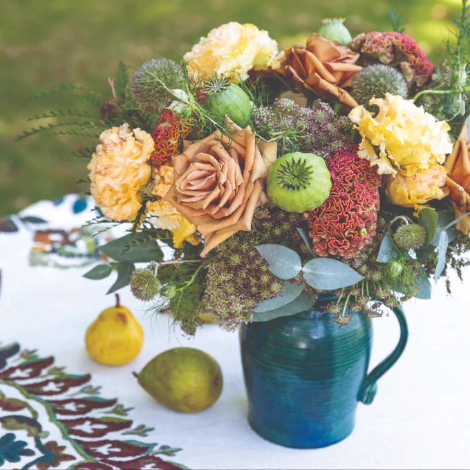 Alt text: A flower arrangement in fall tones of rust, green, cream, and pale orange made of Toffee roses, carnations, celosia, Japanese lisianthus, poppy pods, love-in-a-mist, Queen Anne’s lace, eucalyptus, and euphorbia foliage tucked in a blue stone pitcher. 